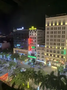 Quang cảnh một góc phố sầm uất vào ban đêm, được chụp từ trên cao. Nhiều tòa nhà cao tầng được chiếu sáng rực rỡ. Dưới đường, xe cộ đang di chuyển và đậu đỗ, cây cối ven đường được trang trí đèn LED.。Các chữ viết và biển hiệu có thể nhận dạng:。- Biển hiệu lớn của khách sạn "DAI VIET HOTEL".。- Biển hiệu của các ngân hàng: "VietinBank", "OCB Ngân Hàng Phương Đông", "eABank", "SeABank".。- Biển hiệu màu vàng của "HOA SEN SPA" với logo hoa sen.。- Một biển hiệu đèn neon màu đỏ chạy dọc có chữ "MASS...", có thể là "MASSAGE".。- Một tấm băng rôn lớn màu đỏ ở góc dưới bên trái có nội dung: "TRUNG TÂM TỔ CHỨC SỰ KIỆN VÀ ẨM THỰC SAO MAI", bên dưới là "TIỆC CƯỚI, HỘI NGHỊ" và "CHUYÊN NGHIỆP".。Hoạt động và vật thể:。- Địa điểm: Một con phố thương mại trong đô thị vào ban đêm.。- Hoạt động: Giao thông đường phố với ô tô và xe máy, người đi bộ trên vỉa hè.。- Vật thể: Các tòa nhà cao tầng (khách sạn, ngân hàng, spa), ô tô, xe máy, cây xanh (bao gồm cả cây cọ), đèn đường, đèn trang trí, các biển quảng cáo và băng rôn.。（日本語訳）。夜の賑やかな街角の一角を上から撮影した光景。多くの高層ビルが煌々とライトアップされている。路上では、車両が移動したり駐車したりしており、沿道の木々はLEDライトで飾られている。認識できる文字と看板：。- ホテル「DAI VIET HOTEL」の大きな看板。- 銀行の看板：「VietinBank」、「OCB Ngân Hàng Phương Đông」、「eABank」、「SeABank」。- 蓮の花のロゴが入った「HOA SEN SPA」の黄色い看板。- 縦に流れる赤いネオンサインに「MASS...」という文字があり、おそらく「MASSAGE」だと思われる。- 左下隅にある大きな赤い横断幕には、「TRUNG TÂM TỔ CHỨC SỰ KIỆN VÀ ẨM THỰC SAO MAI」と書かれており、その下には「TIỆC CƯỚI, HỘI NGHỊ」と「CHUYÊN NGHIỆP」とある。活動と物体：。- 場所：夜の都市の商業的な通り。- 活動：自動車とバイクによる道路交通、歩道上の歩行者。- 物体：高層ビル（ホテル、銀行、スパ）、自動車、バイク、緑樹（ヤシの木を含む）、街灯、装飾灯、広告看板、横断幕。