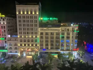 Bức ảnh chụp cảnh một góc phố sầm uất của một thành phố vào ban đêm, được nhìn từ trên cao. Khung cảnh nổi bật với nhiều tòa nhà cao tầng được chiếu sáng rực rỡ bằng đèn trang trí và các biển hiệu đèn neon nhiều màu sắc.。Các dòng chữ trên các biển hiệu được ghi lại như sau:。- Tên khách sạn: "DAI VIET HOTEL", "PALM HOTEL".。- Tên ngân hàng: "ACB" (NGÂN HÀNG Á CHÂU), "OCB" (Ngân Hàng Phương Đông), "eABank", "SeABank".。- Tên các cơ sở kinh doanh khác: "NHÀ SÁCH" (biển hiệu màu đỏ), "...EN SPA" (biển hiệu màu vàng, bị che khuất một phần), "THANH... GROUP" và "Langua..." (biển hiệu màu xanh lá, bị che khuất một phần).。Cảnh vật và hoạt động:。- Địa điểm là một khu phố thương mại với nhiều khách sạn, ngân hàng và cửa hàng.。- Các tòa nhà được trang trí bằng các dải đèn LED chạy dọc theo kiến trúc.。- Dưới mặt đất là một bãi đỗ xe và một con đường có nhiều phương tiện giao thông như ô tô và xe máy đang đậu và di chuyển.。- Có một vài người đang đi bộ trên vỉa hè. Một người đang cầm ô, cho thấy trời có thể đang mưa nhẹ.。- Cây cối dọc theo đường phố cũng được trang trí bằng đèn nháy. Trên sân thượng của một tòa nhà có vẻ là một quán cà phê hoặc quán bar ngoài trời.。（日本語訳）。夜の都市の賑やかな街角を上から撮影した写真です。その光景は、装飾灯や色とりどりのネオンサインで煌々と照らされた多くの高層ビルで際立っています。看板に書かれている文字は以下の通りです。- ホテル名：「DAI VIET HOTEL」、「PALM HOTEL」。- 銀行名：「ACB」（アジア商業銀行）、「OCB」（オリエント商業銀行）、「eABank」、「SeABank」。- その他の事業所名：「NHÀ SÁCH」（書店、赤い看板）、「...EN SPA」（黄色い看板、一部が隠れている）、「THANH... GROUP」および「Langua...」（緑色の看板、一部が隠れている）。風景と活動：。- 場所は、多くのホテル、銀行、店舗が立ち並ぶ商業地区です。- 建物は、建築に沿って走るLEDライトの帯で装飾されています。- 地上には駐車場と、自動車やバイクなどの多くの交通車両が駐車したり移動したりしている道路があります。- 歩道には数人が歩いています。一人が傘をさしており、小雨が降っている可能性を示しています。- 通り沿いの木々もイルミネーションで飾られています。ある建物の屋上には、屋外カフェまたはバーがあるようです。
