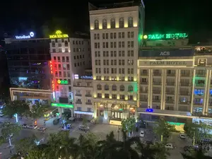 Bức ảnh chụp một góc phố sầm uất của một thành phố vào ban đêm, nhìn từ trên cao. Khung cảnh nổi bật với nhiều tòa nhà cao tầng được chiếu sáng rực rỡ. Có thể nhận dạng rõ các biển hiệu đèn neon của nhiều cơ sở kinh doanh, bao gồm:。- Khách sạn: 'DAI VIET HOTEL', 'PALM HOTEL'.。- Ngân hàng: 'VietinBank', 'ACB', 'eABank', 'OCB' (Ngân Hàng Phương Đông), 'SeABank'.。- Dịch vụ khác: 'HOA SEN SPA', và một biển hiệu dọc màu đỏ có chữ 'MASSAGE'.。Dưới lòng đường và vỉa hè, có nhiều ô tô đang đỗ, một vài người đang đi bộ và có người đang đi xe máy. Một số cây xanh được trang trí bằng đèn nháy, góp phần tạo nên không khí nhộn nhịp của đô thị về đêm.。（日本語訳）。写真は、ある都市の賑やかな街角を夜間に上から撮影したものです。色鮮やかにライトアップされた多くの高層ビルが際立つ光景です。多くの商業施設のネオンサインがはっきりと認識でき、以下が含まれます。- ホテル：「DAI VIET HOTEL」、「PALM HOTEL」。- 銀行：「VietinBank」、「ACB」、「eABank」、「OCB」（東方銀行）、「SeABank」。- その他のサービス：「HOA SEN SPA」、そして「MASSAGE」という文字が入った赤い縦長の看板。車道や歩道には、多くの車が駐車しており、数人が歩いていて、バイクに乗っている人もいます。いくつかの木はイルミネーションで飾られており、夜の都会の賑やかな雰囲気を醸し出しています。