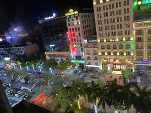 Bức ảnh chụp cảnh một con phố sầm uất vào ban đêm từ trên cao, tại một khu vực đô thị ở thành phố Thanh Hóa. Đường phố và các tòa nhà cao tầng được chiếu sáng rực rỡ bởi đèn đường và các biển hiệu quảng cáo. Có nhiều ô tô đang đỗ dọc hai bên đường và trong bãi đỗ xe, một vài chiếc xe máy và ô tô đang lưu thông. Một số người đang đi bộ trên vỉa hè. Hàng cây xanh, bao gồm cả cây cọ, được trang trí bằng dải đèn LED. Các chữ viết trên biển hiệu có thể nhận dạng được bao gồm: Tên các ngân hàng: VietinBank, OCB (Ngân Hàng Phương Đông), eABank, SeABank, ACB. Tên khách sạn và dịch vụ: DAI VIET HOTEL, HOA SEN SPA, và một phần của biển hiệu 'PAL...'. Tên trung tâm thương mại ở phía xa: VINCOM PLAZA. Một tấm biển quảng cáo lớn màu đỏ ở phía dưới bên trái có nội dung: 'TRUNG TÂM TỔ CHỨC SỰ KIỆN VÀ ẨM THỰC SAO MAI', 'TIỆC CƯỚI, HỘI NGHỊ', 'CHUYÊN NGHIỆP'.。（日本語訳）。タインホア市のある都市部で、夜の賑やかな通りを上から撮影した写真。道路と高層ビルは、街灯や広告看板によって煌々と照らされている。多くの自動車が道路の両脇や駐車場に駐車しており、数台のバイクと自動車が通行している。何人かの人々が歩道を歩いている。ヤシの木を含む緑の並木は、LEDライトの帯で装飾されている。認識できる看板の文字には以下が含まれる：銀行名：VietinBank、OCB（東方銀行）、eABank、SeABank、ACB。ホテルとサービス名：DAI VIET HOTEL、HOA SEN SPA、そして「PAL...」という看板の一部。遠くにあるショッピングセンター名：VINCOM PLAZA。左下にある大きな赤い広告看板には、「SAO MAI イベント・飲食センター」、「結婚披露宴、会議」、「プロフェッショナル」という内容が書かれている。