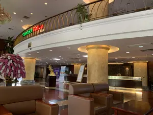 Bối cảnh là sảnh của một khách sạn hoặc tòa nhà lớn, sang trọng. Sàn nhà được đánh bóng, có nhiều cột trụ lớn giả đá cẩm thạch. Ở tiền cảnh là khu vực chờ với bộ ghế sofa và ghế bành bọc da màu nâu. Ở trung tâm sảnh có một bình hoa lớn cắm hoa lan giả màu hồng và trắng, cùng một bức tượng nghệ thuật màu vàng đặt trên bệ. Phía sau là quầy lễ tân dài. Có các tấm biển và poster được dựng lên, trong đó một poster có dòng chữ "CHÀO MỪNG ĐẠI BIỂU THAM DỰ LIÊN HOAN ... ANH TOÀN QUỐC VI - 2024" và một poster khác quảng cáo du lịch với chữ "Sầm Sơn". Nhìn lên tầng trên, có một ban công hình vòng cung với lan can kim loại màu vàng. Trên lan can có một biển hiệu đèn neon với dòng chữ "Grand Bar Coffee" màu xanh lá và đỏ. Không có người xuất hiện trong ảnh, cho thấy một không gian yên tĩnh.。（日本語訳）。背景は、大きくて豪華なホテルまたは建物のロビーです。床は磨かれており、大理石風の大きな柱がたくさんあります。前景には、茶色の革張りのソファとアームチェアのセットがある待合エリアがあります。ロビーの中央には、ピンクと白の造花の蘭が生けられた大きな花瓶と、台座の上に置かれた黄色い芸術的な彫像があります。後方には長いフロントデスクがあります。立てかけられた看板やポスターがあり、そのうちの一つのポスターには「第6回全国...フェスティバルに参加する代表者を歓迎します - 2024」という文言があり、もう一つは「サムソン」という文字で観光を宣伝するポスターです。上の階を見上げると、黄色い金属製の手すりが付いたアーチ型のバルコニーがあります。手すりの上には、緑と赤の「Grand Bar Coffee」という文字のネオンサインがあります。写真には誰も写っておらず、静かな空間を示しています。