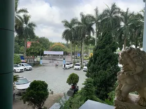 Bức ảnh được chụp từ trên cao, nhìn xuống một khoảng sân hoặc bãi đỗ xe lát gạch. Trong sân có nhiều xe ô tô đang đỗ. Ở phía trước bên phải, có một bức tượng sư tử đá lớn. Khung cảnh được bao quanh bởi nhiều cây xanh, bao gồm các hàng cây cọ cao, cây tùng và cây cảnh. Ở phía xa là một con đường có người đi xe máy và một cổng chào lớn hình vòm. Trên cổng chào có treo một băng rôn trắng không có chữ và dòng chữ trang trí màu vàng nhưng không thể đọc rõ. Có một vài người trong sân; một người đang dắt xe đạp và một người khác đứng cạnh một chiếc ô tô màu trắng đang mở cửa. Có thể nhìn thấy một phần biển quảng cáo màu đỏ với chữ 'GENE...', một biển hiệu nhỏ màu xanh lá cây ghi 'KHÁCH SẠN... MASSAGE', và một biển báo giao thông màu xanh dương nhưng chữ không rõ.。（日本語訳）。写真は上から撮影されており、レンガ敷きの中庭または駐車場を見下ろしています。庭には多くの自動車が駐車しています。右前景には、大きな石のライオンの像があります。風景は、高いヤシの木の列、松の木、観葉植物など、多くの緑の木々に囲まれています。遠くにはバイクに乗った人がいる道路と、大きなアーチ型の歓迎ゲートがあります。歓迎ゲートには、文字のない白い横断幕と、黄色い装飾文字の列が掲げられていますが、はっきりと読むことはできません。庭には数人の人がいます。一人は自転車を押しており、もう一人はドアが開いている白い車の隣に立っています。「GENE...」という文字のある赤い広告看板の一部、「KHÁCH SẠN... MASSAGE（ホテル...マッサージ）」と書かれた小さな緑色の看板、そして文字が不鮮明な青い交通標識が見えます。