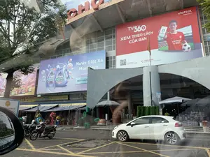 Bức ảnh chụp khung cảnh phía trước một tòa nhà chợ lớn, có thể nhận biết qua chữ "CHỢ" màu đỏ trên nóc nhà. Tòa nhà có nhiều biển quảng cáo lớn. Một biển quảng cáo của "viettel telecom" cho dịch vụ "TV360" với khẩu hiệu "XEM THEO CÁCH CỦA BẠN", có hình một cầu thủ bóng đá và logo "EURO 2024", cùng số điện thoại "1800.8168". Một biển quảng cáo khác dành cho sản phẩm "Nước Giặt Xả MaxKleen" với dòng chữ "SẠCH KHUẨN". Dưới tầng trệt là một dãy các ki-ốt và cửa hàng có mái hiên, đang hoạt động kinh doanh. Các biển hiệu có thể đọc được bao gồm: "ĐẠI LÝ HOA HÀ", "SƠN TRIỆU", "Loan Len CHUYÊN BÁN BUÔN - BÁN LẺ", "LAN TIẾN", "ĐẠI LÝ HOA LỤA HOA SÁP", và "KIM CÚC". Các cửa hàng này bày bán nhiều loại mặt hàng như hoa, đồ gia dụng, thảm, cặp sách. Trên con đường phía trước, có một chiếc ô tô màu trắng và một vài chiếc xe máy đang đỗ. Bức ảnh được chụp từ bên trong một chiếc xe hơi, có thể thấy hình ảnh phản chiếu trên kính cửa sổ và gương chiếu hậu.。（日本語訳）。大きな市場の建物の前の風景を撮影した写真で、屋根の上にある赤い「CHỢ」の文字でそれと分かります。建物には大きな広告看板がいくつもあります。「viettel telecom」の広告看板が一つあり、「TV360」というサービスのもので、「XEM THEO CÁCH CỦA BẠN（あなたのやり方で見る）」というスローガン、サッカー選手の写真、「EURO 2024」のロゴ、そして電話番号「1800.8168」が記載されています。別の広告看板は「Nước Giặt Xả MaxKleen（マックスクリーン洗濯柔軟剤）」という製品のもので、「SẠCH KHUẨN（除菌）」という文字が書かれています。1階には、庇（ひさし）のあるキオスクや店舗が並んでおり、営業しています。読み取れる看板には、「ĐẠI LÝ HOA HÀ」、「SƠN TRIỆU」、「Loan Len CHUYÊN BÁN BUÔN - BÁN LẺ（卸売・小売専門）」、「LAN TIẾN」、「ĐẠI LÝ HOA LỤA HOA SÁP（シルクフラワー・ワックスフラワー代理店）」、そして「KIM CÚC」などがあります。これらの店では、花、家庭用品、カーペット、通学カバンなど、さまざまな種類の商品が陳列販売されています。前の道路には、白い車1台と数台のバイクが駐車しています。写真は車内から撮影されたもので、窓ガラスやバックミラーに反射した像が見えます。