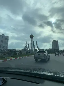 Bức ảnh chụp cảnh quan tại một vòng xuyến giao thông lớn ở trung tâm thành phố Thanh Hóa. Ở giữa vòng xuyến là một tượng đài kiến trúc hiện đại và hoành tráng. Tượng đài này có một cột trụ chính cao, trên đỉnh là một quả cầu lớn có dòng chữ màu vàng, có thể đọc được chữ 'THANH HÓA' và một phần chữ tiếp theo. Xung quanh tượng đài là các công trình điêu khắc bằng bê tông cong vút. Bao quanh vòng xuyến là các tòa nhà cao tầng, trong đó một tòa nhà bên trái có biển hiệu 'Eurowindow Garden City'. Dải phân cách được trồng cây xanh và có lề đường sơn màu đỏ và trắng. Hoạt động giao thông đang diễn ra với nhiều phương tiện. Một chiếc xe bán tải màu bạc hiệu Ford Ranger đang chạy phía trước. Nhiều người đi xe máy cũng đang lưu thông trên đường. Ở phía xa bên trái có một chiếc xe khách màu vàng. Bức ảnh được chụp từ bên trong một chiếc ô tô, nhìn qua kính chắn gió có đọng vài giọt nước, cho thấy thời tiết ẩm ướt hoặc vừa có mưa. Bầu trời u ám và đầy mây.。（日本語訳）。タインホア市中心部にある大きな交通ロータリーの風景を写した写真です。ロータリーの中央には、現代的で壮大な建築のモニュメントがあります。このモニュメントには高い主柱があり、その頂上には大きな球体があり、黄色の文字で「THANH HÓA」という文字とその次の一部の文字が読み取れます。モニュメントの周りには、そびえ立つように湾曲したコンクリートの彫刻作品があります。ロータリーを囲むように高層ビルが建ち並び、そのうち左側のビルには「Eurowindow Garden City」という看板があります。中央分離帯には緑の木が植えられ、縁石は赤と白に塗られています。多くの車両が行き交い、交通が活発です。フォード・レンジャーの銀色のピックアップトラックが前方を走っています。多くのバイクも道路を通行しています。遠く左手には黄色のバスがいます。この写真は車内から撮影されたもので、フロントガラスに水滴がいくつか付着していることから、湿気が多いか、雨が降った直後であることがわかります。空はどんよりと曇っています。