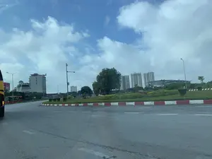 Đây là hình ảnh chụp cảnh quan đô thị tại một vòng xuyến lớn. Con đường nhựa có vạch kẻ đường, và dải phân cách trung tâm được sơn màu đỏ và trắng, bên trên trồng cây xanh và cỏ. Ở phía sau, có nhiều tòa nhà. Một tòa nhà bên trái có biển hiệu ghi chữ "Kol Coffee". Một chiếc xe khách màu đỏ đang ở gần vòng xuyến. Phía xa là một dãy các tòa nhà chung cư cao tầng, hiện đại. Bầu trời xanh và có nhiều mây. Toàn cảnh cho thấy một khu vực đô thị đang phát triển với các phương tiện giao thông đang di chuyển.。（日本語訳）。これは、大きなロータリーの都市景観を撮影した写真です。アスファルトの道路には車線が引かれており、中央分離帯は赤と白に塗られ、その上には緑の木々や草が植えられています。後方には、多くの建物があります。左側にある建物には「Kol Coffee」と書かれた看板があります。赤いバスが1台、ロータリーの近くにいます。遠くには、近代的な高層マンションが立ち並んでいます。空は青く、多くの雲があります。全景は、交通車両が移動している発展中の都市部を示しています。