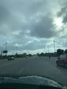 Bức ảnh ghi lại cảnh quan trên một đại lộ rộng lớn có nhiều làn xe ở Thanh Hóa. Khung cảnh cho thấy một con đường thẳng tắp với các phương tiện giao thông như ô tô và xe máy đang di chuyển. Ở bên phải là một chiếc ô tô màu đỏ, bên trái là một chiếc ô tô màu đen, và ở phía xa có nhiều xe cộ khác. Dọc hai bên đường là hàng cột đèn đường cao và cây xanh. Phía xa, con đường dường như dốc lên, có thể dẫn đến một cây cầu. Bầu trời u ám, đầy mây, nhưng vẫn có ánh nắng xuyên qua. Có một số biển báo giao thông, bao gồm một biển báo lớn phía trên cao ở bên trái, nhưng nội dung không thể đọc được. Bức ảnh được chụp từ góc nhìn bên trong một chiếc xe ô tô, với những giọt nước còn đọng lại trên kính chắn gió ở phía dưới.。（日本語訳）。写真は、タインホア省にある複数の車線を持つ広大な大通りの景観を記録したものです。その光景は、自動車やバイクなどの交通車両が移動している、まっすぐな道を示しています。右側には赤い自動車が、左側には黒い自動車が、そして遠くには他の多くの車両が見えます。道路の両脇には、高い街灯の列と緑の木々が並んでいます。遠くでは、道は上り坂になっているようで、橋につながっている可能性があります。空はどんよりとして雲に覆われていますが、それでも日差しが差し込んでいます。いくつかの交通標識があり、左側の上方には大きな標識がありますが、その内容は読み取れません。写真は自動車の中から撮影されたもので、下部のフロントガラスには水滴が残っています。