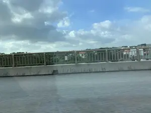 Bức ảnh được chụp từ một phương tiện đang di chuyển trên một cây cầu hoặc đường trên cao. Phía trước là mặt đường nhựa bị mờ do chuyển động và một lan can cầu bằng bê tông với tay vịn kim loại. Nhìn qua lan can là khung cảnh một khu dân cư với nhiều nhà cửa và cây cối xanh tươi. Bầu trời có nhiều mây trắng và xám xen lẫn những khoảng trời xanh. Không có văn bản hay chữ viết nào trong ảnh.。（日本語訳）。写真は、橋または高架道路の上を移動中の乗り物から撮影されたものです。前景には、動きによってぼやけたアスファルトの路面と、金属製の手すりが付いたコンクリート製の橋の欄干があります。欄干の向こうには、多くの家々と青々とした木々がある住宅街の風景が広がっています。空には白と灰色の雲が多く、青空が混じっています。写真の中には、いかなるテキストや文字もありません。