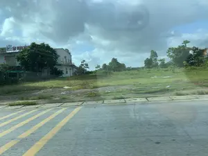 Bức ảnh chụp một khung cảnh ven đường từ một phương tiện đang di chuyển. Ở phía trước là mặt đường nhựa với các vạch kẻ chéo màu vàng. Bên cạnh đường là một khu đất trống, cỏ mọc um tùm. Ở phía sau là một tòa nhà màu trắng nhiều tầng, có treo cờ Việt Nam. Trên nóc tòa nhà có một biển hiệu lớn màu đỏ với dòng chữ '36 TẬP ĐOÀN ĐẦU...', phần còn lại bị cây che khuất. Một biển hiệu nhỏ hơn có chữ 'ROCKSTAR' cũng có thể được nhìn thấy. Xung quanh tòa nhà và khu đất có nhiều cây xanh. Bầu trời có nhiều mây.。（日本語訳）。移動中の乗り物から撮影された道端の風景写真です。手前には、黄色い斜線が引かれたアスファルトの路面があります。道路の隣には、草が鬱蒼と茂る空き地があります。奥には、ベトナム国旗が掲げられた白い多層階の建物があります。建物の屋上には、「36 TẬP ĐOÀN ĐẦU...」という文字が書かれた大きな赤い看板があり、残りの部分は木で隠されています。「ROCKSTAR」という文字が入った小さな看板も見えます。建物と空き地の周りには、たくさんの緑の木々があります。空は雲が多いです。