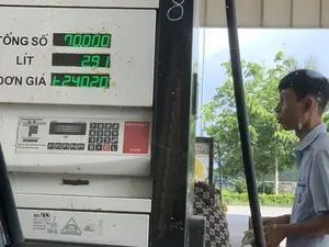 Bối cảnh là một trạm xăng. Ở cận cảnh là một trụ bơm xăng đã có dấu hiệu cũ và gỉ sét. Trên màn hình điện tử của trụ bơm hiển thị các thông số của một giao dịch: 'TỔNG SỐ' là 70000 (đồng), 'LÍT' là 2.91, và 'ĐƠN GIÁ' là 24020 (đồng/lít). Bên dưới màn hình là bàn phím số và các hướng dẫn sử dụng bằng tiếng Anh, bao gồm 'Preset instructions' và 'EMERGENCY STOP'. Bên phải là một nhân viên trạm xăng, là một người đàn ông mặc đồng phục màu xanh nhạt, đang đứng cạnh trụ bơm. Phía sau là khung cảnh ngoài trời với cây cối xanh tươi.。（日本語訳）。背景はガソリンスタンドです。クローズアップには、古びて錆びついた給油機があります。給油機の電子ディスプレイには、ある取引の各数値が表示されています。「合計金額」は70000（ドン）、「リットル」は2.91、「単価」は24020（ドン/リットル）です。ディスプレイの下には、数字キーパッドと、「Preset instructions」や「EMERGENCY STOP」といった英語の使用説明があります。右側には、水色の制服を着た男性のガソリンスタンド従業員が給油機の隣に立っています。後ろには、青々とした木々のある屋外の風景が広がっています。