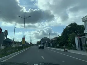 Bức ảnh chụp khung cảnh một con đường nhựa rộng ở Việt Nam, được chụp từ góc nhìn của một người đang ngồi trong ô tô. Bầu trời có nhiều mây xám, cho thấy một ngày trời râm mát. Hai bên đường là các ngôi nhà dân sinh, cây cối và cột điện. Một chiếc ô tô màu đen đang di chuyển ở phía trước. Bên lề đường phải, một người đội nón lá đang ngồi trên một chiếc xe máy. Ở phía bên trái, có một tấm phướn tuyên truyền màu đỏ và vàng treo trên cột, trên đó có dòng chữ: "GIỮ GÌN VÀ BẢO VỆ CÁC DI TÍCH VĂN HÓA LỊCH SỬ LÀ TRÁCH NHIỆM CỦA MỖI NGƯỜI DÂN。". Phía dưới là logo của ngân hàng "BIDV".。（日本語訳）。ベトナムの広いアスファルト道路の風景を写した写真で、車の中に座っている人の視点から撮影されたものです。空は灰色の雲が多く、曇りの涼しい日であることがわかります。道路の両側には民家、木々、電柱があります。前方を黒い車が走行しています。右側の路肩では、ノンラーをかぶった人がバイクに座っています。左側には、赤と黄色の宣伝用の垂れ幕が柱に掲げられており、そこには「歴史文化遺産を維持・保護することは、国民一人ひとりの責任です。」という文言が書かれています。その下には「BIDV」銀行のロゴがあります。