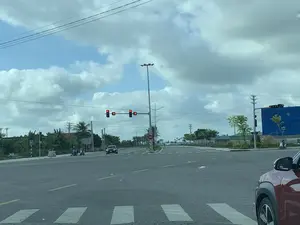 Bức ảnh chụp một ngã tư đường phố rộng rãi vào ban ngày, dưới bầu trời xanh có nhiều mây. Tại ngã tư, có hệ thống đèn tín hiệu giao thông đang bật màu đỏ, kèm theo đồng hồ đếm ngược hiển thị số '8'. Một chiếc ô tô màu đen và một vài chiếc xe máy đang dừng lại chờ đèn xanh. Ở góc dưới bên phải, có thể thấy một phần của một chiếc ô tô màu đỏ. Con đường có vạch kẻ đường rõ ràng, bao gồm cả vạch dành cho người đi bộ. Hai bên đường được trồng cây xanh, có các cột đèn chiếu sáng và dây điện. Phía xa bên phải, một công trình xây dựng được che phủ bởi một tấm bạt lớn màu xanh. Khung cảnh cho thấy các phương tiện đang tham gia giao thông và tuân thủ tín hiệu đèn đỏ.。（日本語訳）。日中の広々とした交差点を写した写真で、空は青く、多くの雲が浮かんでいます。交差点では、交通信号機が赤色に点灯しており、「8」という数字を表示するカウントダウンタイマーが付いています。一台の黒い車と数台のバイクが青信号を待って停止しています。右下隅には、赤い車の一部が見えます。道路には、横断歩道を含む明確な車線が引かれています。道路の両側には街路樹が植えられ、街灯や電線があります。右手奥には、大きな青いシートで覆われた建設現場があります。この光景は、車両が交通に参加し、赤信号の指示に従っていることを示しています。