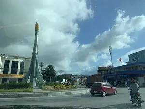 Đây là hình ảnh chụp một vòng xuyến giao thông tại một khu vực đô thị, có thể là Thị trấn Bút Sơn, tỉnh Thanh Hóa. Ở trung tâm vòng xuyến có một công trình điêu khắc hiện đại, cao, trông giống một ngọn đuốc hoặc một cây bút chì khổng lồ. Xung quanh vòng xuyến có nhiều tòa nhà và biển hiệu. Bên trái là tòa nhà Bưu điện Việt Nam với biển hiệu 'BƯU ĐIỆN VIỆT NAM' và logo 'VIETNAM POST'. Bên phải là một trạm xăng PVOIL với biển hiệu 'CỬA HÀNG XĂNG DẦU SỐ 1 THỊ TRẤN BÚT SƠN' và lời chào 'Kính Chào Quý Khách。'. Trên nóc trạm xăng có một lá cờ Việt Nam và một biển quảng cáo lớn màu xanh ghi 'Chống thấm TƯỜNG & SÀN thế hệ mới'. Các biển hiệu khác như 'BÁN TRẢ GÓP' và 'WinMart' cũng có thể được nhìn thấy ở phía xa. Hoạt động chính là giao thông đường bộ với các phương tiện như ô tô, xe máy và xe tải container đang di chuyển. Bầu trời có mây xanh.。（日本語訳）。これは、ある都市部の交通ロータリーを撮影した写真で、おそらくタインホア省ブットソン町でしょう。ロータリーの周りには、多くの建物や看板があります。左側には、「BƯU ĐIỆN VIỆT NAM」という看板と「VIETNAM POST」のロゴがあるベトナム郵便の建物があります。右側には、「CỬA HÀNG XĂNG DẦU SỐ 1 THỊ TRẤN BÚT SƠN」（ブットソン町第1号ガソリンスタンド）という看板と「Kính Chào Quý Khách。」（お客様、こんにちは。）という挨拶があるPVOILのガソリンスタンドがあります。ガソリンスタンドの屋根の上には、ベトナム国旗と、「Chống thấm TƯỜNG & SÀN thế hệ mới」（新世代の壁＆床用防水）と書かれた大きな青い広告看板があります。「BÁN TRẢ GÓP」（分割払い販売）や「WinMart」といった他の看板も遠くに見えます。主な活動は道路交通で、自動車、バイク、コンテナトラックなどの車両が移動しています。空は青く、雲があります。