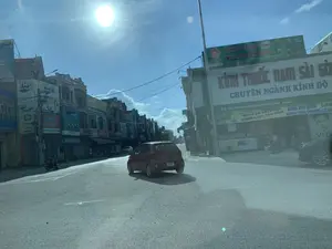 Bối cảnh là một con đường tại một khu vực đô thị ở Việt Nam vào một ngày nắng. Hình ảnh được chụp từ bên trong một chiếc xe hơi, nhìn ra một ngã tư hoặc một đoạn đường đông đúc. Một chiếc ô tô nhỏ màu đỏ đang chạy trên đường. Hai bên đường là những dãy nhà phố liền kề, nhiều tầng, với kiến trúc đa dạng. Các tòa nhà này được sử dụng làm mặt bằng kinh doanh với rất nhiều biển hiệu quảng cáo. Bên phải, có một cửa hàng lớn với biển hiệu "KÍNH THUỐC NAM SÀI GÒN - CHUYÊN NGÀNH KÍNH ĐO", địa chỉ được ghi rõ là "CS1: SN 103 - 105 Phố Trung Sơn vòng xoay ngã 5 Bút Sơn - Hoằng Hóa - Thanh Hóa". Cửa hàng này có các băng rôn quảng cáo các chương trình như "CHÀO HÈ SÔI ĐỘNG", "KÍNH ĐỔI MÀU", "KÍNH THỜI TRANG" và dịch vụ "KHÚC XẠ MIỄN PHÍ". Phía trên cửa hàng kính là một biển hiệu màu xanh lá cây có chữ "MUA CHÁO TẶNG THẺ QUÀ TÍCH ĐIỂM". Dãy nhà bên trái có nhiều biển hiệu khác nhau, bao gồm "Tiền Có Ngay" (dịch vụ tài chính), "oppo" (thương hiệu điện thoại), "BIA HÀ NỘI", "MINH SÂM". Một người đang điều khiển xe máy cũng xuất hiện ở phía bên trái đường. Khung cảnh thể hiện hoạt động giao thông và thương mại thường ngày.。（日本語訳）。背景は、晴れた日のベトナムの都市部にある道路です。写真は車の中から撮影されたもので、交差点か混雑した道路が見えます。一台の小さな赤い車が道路を走っています。道路の両側には、多様な建築様式の多層階建てのタウンハウスが立ち並んでいます。これらの建物は、たくさんの広告看板がある商業施設として使用されています。右側には、「KÍNH THUỐC NAM SÀI GÒN - CHUYÊN NGÀNH KÍNH ĐO」（サイゴン南薬眼鏡店 - 眼鏡測定専門）という看板を掲げた大きな店があり、住所は「CS1: SN 103 - 105 Phố Trung Sơn vòng xoay ngã 5 Bút Sơn - Hoằng Hóa - Thanh Hóa」（1号店：タインホア省ホアンホア県ブットソン5差路ロータリー、チュンソン通り103-105番地）と明記されています。この店には、「CHÀO HÈ SÔI ĐỘNG」（エキサイティングな夏を迎えよう）、「KÍNH ĐỔI MÀU」（調光レンズ）、「KÍNH THỜI TRANG」（ファッションメガネ）といったキャンペーンや、「KHÚC XẠ MIỄN PHÍ」（無料屈折検査）というサービスを宣伝する横断幕があります。眼鏡店の上には、「MUA CHÁO TẶNG THẺ QUÀ TÍCH ĐIỂM」（お粥を買ってポイントカードをプレゼント）と書かれた緑色の看板があります。左側の建物群には、「Tiền Có Ngay」（即日融資、金融サービス）、「oppo」（携帯電話ブランド）、「BIA HÀ NỘI」（ハノイビール）、「MINH SÂM」など、さまざまな看板があります。この光景は、日常の交通と商業活動を表しています。