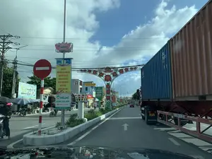 Bức ảnh chụp cảnh đường phố tại huyện Hoằng Hóa, tỉnh Thanh Hóa, nhìn từ trong một chiếc ô tô. Con đường có nhiều phương tiện giao thông đang di chuyển, bao gồm một chiếc xe tải container lớn màu xanh và nâu ở bên phải, một chiếc ô tô màu đỏ ở phía xa và nhiều xe máy. Con đường có một cổng chào trang trí với họa tiết hoa màu đỏ. Dọc theo dải phân cách và hai bên đường có nhiều biển báo và băng rôn quảng cáo. Có một biển báo giao thông hình tròn màu đỏ cấm đi ngược chiều. Các văn bản có thể đọc được trên các biểu ngữ và biển hiệu bao gồm:。- Băng rôn tuyên truyền: 'ĐẢNG BỘ VÀ NHÂN DÂN HUYỆN HOẰNG HÓA QUYẾT TÂM THỰC HIỆN THẮNG LỢI NGHỊ QUYẾT ĐẠI HỘI ĐẢNG BỘ HUYỆN LẦN THỨ XXVII, NHIỆM KỲ 2020-2025。'.。- Biển quảng cáo y tế: 'HỆ THỐNG Y TẾ C+ MEDIC', 'MEDIC HẢI TIẾN', số điện thoại '0915.13.15.15' và khẩu hiệu 'CÙNG HOẰNG HÓA VƯƠN XA'.。- Biển hiệu cửa hàng: 'QUANG SAU MUA BÁN, KÝ GỬI XE MÁY', 'BẤT ĐỘNG SẢN MEDILAND 0849.75.78.75', và 'WinMart'.。- Một biển quảng cáo hình hoa sen của 'C+ MEDIC'.。Bên lề đường là các tòa nhà, cửa hàng và cây cối. Một số người dân đang điều khiển xe máy.。（日本語訳）。タインホア省ホアンホア県の路上風景を、一台の車内から撮影した写真。道路には多くの交通車両が移動しており、右側には青と茶色の大きなコンテナトラック1台、遠くには赤い乗用車1台、そして多くのバイクが含まれている。道路には赤い花の模様で装飾された歓迎ゲートがある。中央分離帯と道路の両側には、多くの標識や広告バナーがある。逆走を禁止する赤い円形の交通標識がある。バナーや看板で読み取れるテキストは以下の通り：。- 宣伝バナー：「ホアンホア県党委員会と人民は、2020-2025年任期、第27回県党大会決議の勝利的な実現を決意する。」。- 医療広告看板：「C+ MEDIC医療システム」、「MEDICハイティエン」、電話番号「0915.13.15.15」、スローガン「ホアンホアと共に遠くへ」。- 店舗の看板：「QUANG SAU バイク売買・委託販売」、「MEDILAND不動産 0849.75.78.75」、そして「WinMart」。- 「C+ MEDIC」の蓮の花の形をした広告看板。道端には建物、店、そして木々がある。何人かの人々がバイクを運転している。