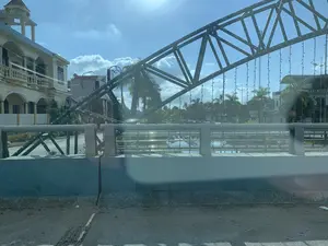 Bức ảnh ghi lại khung cảnh trên một cây cầu, có thể được chụp từ bên trong một phương tiện đang di chuyển. Điểm nổi bật trong ảnh là một kết cấu giàn thép lớn, được trang trí bằng nhiều dây đèn LED, đã bị đổ nghiêng và gác lên lan can cầu. Tình trạng này cho thấy có thể đã xảy ra một sự cố. Bên dưới cầu là một con sông hoặc kênh, mặt nước phản chiếu ánh nắng. Ở phía bên kia bờ là các công trình nhà ở, trong đó có một ngôi nhà lớn mang kiến trúc kiểu biệt thự, và nhiều cây xanh như cây cọ. Bầu trời trong xanh, có nắng và mây trắng. Không có văn bản hay biển hiệu nào có thể đọc được. Một chi tiết nhỏ là có một vật giống ấm trà bằng kim loại đặt trên dải phân cách của cầu.。（日本語訳）。写真は、ある橋の上の光景を記録したもので、移動中の乗り物の中から撮影された可能性があります。写真の中で際立っているのは、多くのLEDライトストリングで装飾された大きな鋼鉄のトラス構造物で、傾いて倒れ、橋の欄干に寄りかかっています。この状況は、何らかの事故が起こった可能性を示しています。橋の下は川か運河で、水面は日光を反射しています。対岸には住宅建築物があり、その中にはヴィラ風の建築様式の大きな家や、ヤシの木のような多くの緑の木々があります。空は青く澄んでおり、晴れていて白い雲があります。読み取れるテキストや看板はありません。小さなディテールとして、橋の中央分離帯の上に金属製のティーポットのような物が置かれています。