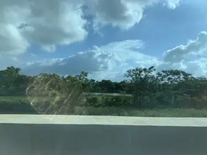 Bức ảnh chụp một khung cảnh thiên nhiên, có thể được chụp từ trên một cây cầu hoặc một con đường. Ở phía trước là một lan can bằng bê tông. Phía sau lan can là một con sông hoặc hồ nước, được bao quanh bởi cây cối và thảm thực vật xanh tươi, rậm rạp. Bầu trời trong xanh, có nhiều mây trắng. Không có bất kỳ văn bản, biển báo hay hoạt động cụ thể nào của con người được nhìn thấy trong ảnh.。（日本語訳）。写真は自然の風景を写したもので、橋の上か道路から撮影された可能性があります。手前にはコンクリート製の手すりがあります。手すりの後ろには川か湖があり、青々とした鬱蒼とした木々や植物に囲まれています。空は青く澄んでおり、白い雲が多く浮かんでいます。写真には、いかなる文字、標識、または特定の人の活動も見られません。