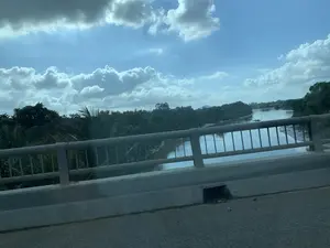 Bức ảnh được chụp từ trên một cây cầu, nhìn ra khung cảnh một con sông ở vùng nông thôn. Ở tiền cảnh là lan can cầu bằng kim loại và thành cầu bê tông. Dưới cầu là một con sông rộng, nước màu nâu đục. Hai bên bờ sông được bao phủ bởi cây cối xanh um tùm, trong đó có cả những cây dừa. Xa xa trên mặt nước, có một chiếc thuyền nhỏ, dường như có người đang hoạt động trên đó, có thể là đánh bắt cá hoặc di chuyển trên sông. Bầu trời phía trên có mây trắng và xám xen lẫn trên nền trời xanh.。（日本語訳）。写真は橋の上から撮影され、田舎にある川の風景を見渡しています。前景には、金属製の橋の欄干とコンクリートの橋の壁があります。橋の下には広い川があり、水は濁った茶色をしています。川の両岸は青々と生い茂る木々に覆われており、その中にはヤシの木もあります。遠くの水面には小さな船が一隻あり、その上では人が活動しているようです。それは漁をしているか、あるいは川を移動しているのかもしれません。上空には、青空を背景に白と灰色の雲が混じり合っています。