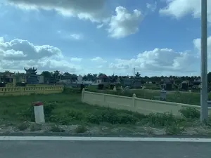 Hình ảnh chụp một khu nghĩa trang rộng lớn nhìn từ một con đường. Bầu trời trong xanh, có nhiều mây trắng. Khu nghĩa trang được bao quanh bởi một bức tường thấp màu vàng. Bên trong có rất nhiều ngôi mộ và lăng mộ được xây dựng công phu với nhiều kiểu kiến trúc khác nhau, một số có mái cong theo phong cách truyền thống. Các ngôi mộ được làm từ đá với các màu sắc như xám, đen và trắng. Cảnh quan xung quanh là đồng cỏ và cây cối. Ở rìa đường phía trước có một cột mốc lộ giới (cọc tiêu). Không có hoạt động nào của con người diễn ra và không có văn bản nào có thể đọc được trong ảnh.。（日本語訳）。ある道から見た広大な墓地の写真。空は澄んだ青色で、白い雲がたくさん浮かんでいる。墓地は黄色い低い壁で囲まれている。内部には、様々な建築様式で精巧に建てられた多くの墓や廟があり、中には伝統的な様式の曲線的な屋根を持つものもある。墓は灰色、黒、白などの色の石でできている。周囲の景観は草原と木々である。手前の道路の端には、道路境界標（デリニエーターポスト）が1本ある。人間の活動は見られず、写真の中に読み取れる文字もない。