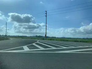 Bức ảnh chụp một con đường nhựa ở khu vực nông thôn vào một ngày trời trong xanh. Bầu trời xanh biếc với những đám mây trắng bồng bềnh. Ở phía trước là một đoạn đường rẽ có vạch kẻ đường hình xương cá màu trắng nổi bật trên nền đường nhựa màu xám. Hai bên đường là những cánh đồng lúa xanh mướt trải dài. Có những cột điện và đường dây điện chạy dọc theo con đường. Ở phía xa, có thể thấy thấp thoáng nhà cửa, cây cối, một công trình trông giống nhà thờ với tháp chuông và bóng dáng của những ngọn núi. Có một tấm biển màu vàng và đỏ gắn trên cột điện nhưng chữ viết không thể đọc được. Khung cảnh yên bình, không có người hay phương tiện giao thông.。（日本語訳）。青空が広がる晴れた日に、田舎のアスファルトの道を撮影した写真です。空は紺碧で、白い雲がふわりと浮かんでいます。手前には分岐路があり、灰色のアスファルト路面に白いヘリンボーン（魚の骨）模様の路面標示が際立っています。道の両側には、青々とした水田が広がっています。道に沿って電柱と電線が走っています。遠くには、家々、木々、鐘楼のある教会のような建物、そして山々の影がかすかに見えます。電柱に黄色と赤色の看板が取り付けられていますが、文字は読み取れません。人や車はおらず、穏やかな光景です。