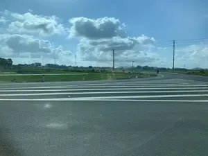 Hình ảnh chụp một con đường nhựa ở vùng nông thôn vào một ngày nắng đẹp. Bầu trời trong xanh và có nhiều mây trắng. Con đường có các vạch kẻ giảm tốc hoặc vạch cho người đi bộ được sơn màu trắng. Bên cạnh đường là một cánh đồng lúa xanh. Dọc theo con đường có các cột điện và cọc tiêu. Ở phía xa, có thể nhìn thấy một vài ngôi nhà, cây cối và những dãy núi. Có một người đang đi bộ và một người khác đang đi xe đạp trên đường.。（日本語訳）。ある晴れた日の田舎のアスファルト道路を撮影した写真です。空は青く澄み渡り、白い雲がたくさん浮かんでいます。道路には、減速帯または横断歩道を示す白い線が引かれています。道の脇には緑の田んぼが広がっています。道に沿って電柱とデリニエーター（視線誘導標）があります。遠くには、いくつかの家、木々、そして山々が見えます。道路上を歩いている人が一人と、自転車に乗っている人がもう一人います。