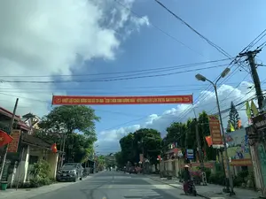 背景はベトナムのある住宅街の道で、おそらくタインホア省ハウロック県、雲のある青空の下です。道を横切って、黄色い文字で「ハウロック県第7回『模範的退役軍人』競走大会（2019年～2024年期）を熱烈に歓迎します」と書かれた大きな赤い横断幕が掲げられています。道の両側には民家、木々、そして多くの電線が張られた電柱があります。多くの国旗と幟旗が道沿いに掲げられています。右側には、ノンラーをかぶった女性が「バス停」の標識の近くでバイクのそばに立っています。その近くにはプロパガンダの壁画や、「ベトナム社会主義共和国万歳。」や「…新しい農村を建設する」といった内容の他の横断幕があります。路上には自動車やバイクなどのいくつかの乗り物が通行しています。この光景は、日常生活の中で政治社会的なイベントが行われている雰囲気を示しています