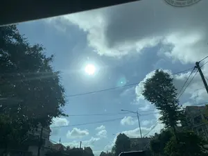 Bức ảnh được chụp từ bên trong một chiếc ô tô, nhìn lên bầu trời vào một ngày nắng. Bầu trời có màu xanh lam với những đám mây trắng và xám rải rác. Mặt trời chiếu sáng mạnh, gây ra hiện tượng lóa ống kính. Khung cảnh hai bên là những tán cây xanh và một phần các tòa nhà, nhà ở ven đường. Có thể thấy rõ hệ thống cột điện, cột đèn đường và dây điện chằng chịt. Ở góc trên của ảnh, có một phần nội thất của xe ô tô và một nhãn dán hình tròn trên kính chắn gió nhưng chữ trên đó không thể đọc được.。（日本語訳）。写真は、ある晴れた日に車の中から空を見上げて撮影されたものです。空は青く、白と灰色の雲が点在しています。太陽が強く輝いており、レンズフレア現象を引き起こしています。両側の風景は、緑の木々の梢と、道端の建物や家の一部です。電柱、街灯柱、そして入り組んだ電線のシステムがはっきりと見えます。写真の上隅には、車内の一部とフロントガラスに貼られた丸いステッカーがありますが、その上の文字は読み取れません。