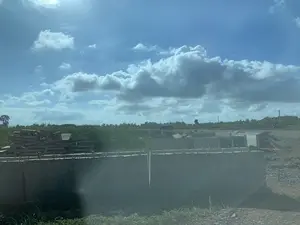 Bức ảnh chụp một khung cảnh ngoài trời, dường như là một công trường xây dựng hoặc một bãi tập kết vật liệu. Ở phía trước và trung tâm bức ảnh là các khối bê tông lớn và các tấm bê tông đúc sẵn, một số được xếp chồng lên nhau. Phía sau các khối bê tông là một khu vực có cỏ dại mọc cao. Ở phía xa là hàng cây xanh và các cột điện dưới bầu trời xanh có nhiều mây trắng. Không có văn bản hay chữ viết nào có thể nhận dạng được trong ảnh. Khung cảnh tĩnh lặng, không có hoạt động nào của con người đang diễn ra.。（日本語訳）。写真は屋外の風景を写しており、建設現場か資材置き場のようです。写真の手前と中央には、大きなコンクリートブロックとプレキャストコンクリート板があり、いくつかは積み重ねられています。遠くには、白い雲が多く浮かぶ青空の下に、緑の木々の列と電柱が見えます。写真の中には、認識できるテキストや文字はありません。風景は静かで、人間の活動は見られません。