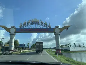 Bức ảnh chụp cảnh quan một con đường ở Việt Nam, được chụp từ góc nhìn bên trong một chiếc ô tô. Điểm nổi bật nhất là một cổng chào lớn, hoành tráng bắc ngang qua đường. Trên thanh ngang của cổng có dòng chữ màu đỏ 'CHÚC QUÝ KHÁCH THƯỢNG LỘ BÌNH AN。'. Phần đỉnh của cổng được trang trí bằng các họa tiết mang đậm nét văn hóa Việt Nam như hình ảnh trống đồng Đông Sơn ở trung tâm và các hình chim Lạc cách điệu.。Một chiếc xe tải đang di chuyển trên đường, đi qua dưới cổng chào. Con đường được trải nhựa và có vạch kẻ. Hai bên đường là khung cảnh làng quê. Bên phải là một ao nước, phía sau là một bức tường trắng của một công trình xây dựng. Bên trái có một vài cửa hàng và các biển quảng cáo, trong đó có biển hiệu 'XÂY DỰNG LINH HOÀNG KIÊM XE CƠ GIỚI 36-10D Thanh Hóa' và một biển hiệu khác có chữ 'BIA HƠI TAM AN'. Bầu trời trong xanh với những đám mây trắng, báo hiệu một ngày thời tiết đẹp.。（日本語訳）。ベトナムのある道路の風景を、車内からの視点で撮影した写真です。最も目立つのは、道路をまたぐように建てられた、大きくて壮大な歓迎の門です。門の横梁には、「CHÚC QUÝ KHÁCH THƯỢNG LỘ BÌNH AN。」という赤い文字があります。門の頂上部分は、中央にドンソン銅鼓のイメージ、そして様式化されたラック鳥の姿など、ベトナム文化の特色が色濃く反映された模様で装飾されています。一台のトラックが道路を走行しており、歓迎の門の下を通過しています。道路は舗装されており、車線が引かれています。道路の両側には田舎の風景が広がっています。右側には池があり、その後ろには建設中の建物の白い壁が見えます。左側にはいくつかの店や広告看板があり、その中には「XÂY DỰNG LINH HOÀNG KIÊM XE CƠ GIỚI 36-10D Thanh Hóa」という看板や、「BIA HƠI TAM AN」という文字のある別の看板があります。空は青く澄み渡り、白い雲が浮かんでおり、天気の良い日であることを示しています。