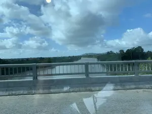 Bức ảnh được chụp từ một phương tiện đang di chuyển khi đi qua một cây cầu. Nhìn qua lan can cầu bằng kim loại, có thể thấy một con sông rộng lớn với dòng nước màu nâu đục. Hai bên bờ sông là thảm thực vật xanh tươi và cây cối rậm rạp. Ở phía xa, có thể nhìn thấy một vài ngôi nhà của khu dân cư và những ngọn đồi. Bầu trời trong xanh với nhiều đám mây trắng và xám. Có một vật thể giống như phao tiêu hàng hải trên mặt sông. Không có hoạt động rõ ràng của con người trên sông, nhưng có vẻ có một vài chiếc thuyền nhỏ neo đậu gần bờ. Không có văn bản hay biển báo nào có thể nhận dạng được trong ảnh.。（日本語訳）。写真は、橋を渡る際に移動中の乗り物から撮影されたものです。金属製の橋の欄干越しに、茶色く濁った水が流れる大きな川が見えます。川の両岸は、青々とした植生と生い茂る木々で覆われています。遠方には、住宅地の家が数軒と丘が見えます。空は青く、白と灰色の雲が多く浮かんでいます。川面には航路標識のような物体があります。川の上ではっきりとした人の活動は見られませんが、岸の近くに数隻の小さなボートが停泊しているようです。写真には、識別できる文字や標識はありません。