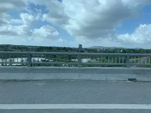 Bức ảnh được chụp từ một phương tiện đang di chuyển, nhìn ra ngoài từ một cây cầu. Phía trước là lan can cầu bằng bê tông và kim loại, bên dưới là mặt đường nhựa. Nhìn qua lan can là một khung cảnh nông thôn với một vùng nước rộng, có thể là một con sông hoặc các ao, đầm nuôi trồng thủy sản. Xung quanh vùng nước là cây cối xanh tươi, bao gồm cả cây chuối. Ở phía xa là những ngôi nhà của người dân nằm rải rác và những dãy núi thấp ở đường chân trời. Bầu trời trong xanh và có nhiều mây trắng. Không có chữ viết hay biển báo nào có thể đọc được trong ảnh.。（日本語訳）。写真は移動中の乗り物から、橋の上から外を眺めるように撮影されたものです。前方にはコンクリートと金属でできた橋の欄干があり、その下はアスファルトの路面です。欄干の向こうには広大な水域のある田園風景が広がっており、それは川であるか、あるいは養殖用の池や沼かもしれません。水域の周りには青々とした木々があり、バナナの木も含まれています。遠くには民家が点在し、地平線には低い山々が連なっています。空は青く澄み渡り、白い雲が多く浮かんでいます。写真の中には読み取れる文字や標識はありません。