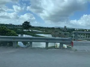 Bức ảnh được chụp từ trên một cây cầu hoặc đường trên cao, nhìn ra khung cảnh một vùng nông thôn ở Việt Nam. Ở phía trước là lan can cầu bằng kim loại. Phía sau lan can là một khu vực rộng lớn với nhiều ao, hồ được chia thành các ô để nuôi trồng thủy sản. Xen kẽ giữa các ao là những ngôi nhà dân, cây cối xanh tươi và các bờ đất. Ở phía xa có thể nhìn thấy nhiều nhà cửa hơn và một dãy núi. Bầu trời trong xanh và có nhiều mây trắng. Không có hoạt động cụ thể nào của con người được ghi lại, nhưng cảnh quan cho thấy đây là một khu vực chuyên về nông nghiệp và nuôi trồng thủy sản. Không có chữ viết nào có thể đọc được trong ảnh.。（日本語訳）。写真は橋または高架道路の上から撮影されたもので、ベトナムの田園地帯の風景を見渡しています。手前には金属製の手すりがあります。手すりの後ろには、水産養殖のために区画に分けられた多くの池や湖がある広大なエリアが広がっています。池の間には、民家、青々とした木々、そして土手が点在しています。遠くには、より多くの家々と山脈が見えます。空は青く澄んでおり、白い雲が多く浮かんでいます。特定の人間の活動は記録されていませんが、風景はここが農業と水産養殖を専門とする地域であることを示しています。写真には読み取れる文字はありません。