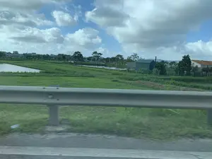 Bức ảnh được chụp từ một phương tiện đang di chuyển, nhìn ra khung cảnh nông thôn qua lan can bảo vệ ven đường. Phong cảnh bao gồm các cánh đồng xanh mướt, có thể là ruộng lúa, và các ao hồ dùng cho việc nuôi trồng thủy sản. Ở phía xa, có thể thấy những ngôi nhà dân, cây cối và một công trình lợp tôn màu xanh. Bầu trời trong xanh, có nhiều mây trắng, tạo nên một khung cảnh yên bình đặc trưng của vùng quê. Không có hoạt động cụ thể nào của con người được ghi lại, nhưng cảnh vật cho thấy đây là một khu vực sản xuất nông nghiệp và nuôi trồng thủy sản.。（日本語訳）。写真は移動中の乗り物から、道路脇のガードレール越しに田園風景を見て撮影されたものです。風景には、青々とした畑（おそらく水田）と、水産養殖に使われる池が含まれています。遠くには、民家、木々、そして青いトタン屋根の建物が見えます。空は青く澄み、白い雲が多く、田舎特有の平和な光景を作り出しています。人間の具体的な活動は記録されていませんが、景色はここが農業生産と水産養殖の地域であることを示しています。