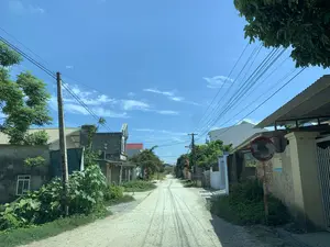 Bức ảnh chụp một con đường làng ở vùng nông thôn vào một ngày nắng. Con đường là đường đất, có nhiều vết bánh xe. Hai bên đường là những ngôi nhà dân với kiến trúc đơn giản, một số có mái tôn, một số có mái ngói đỏ. Cây cối xanh tốt mọc dọc hai bên đường. Hệ thống dây điện, dây cáp chằng chịt vắt ngang qua các cột điện. Ở phía bên phải, có một chiếc gương lồi cầu giao thông. Trên bức tường bên dưới gương có dán một biển quảng cáo nhỏ màu xanh có thể đọc được chữ "INTERNET TRUYỀN HÌNH" và một số điện thoại không rõ. Ngoài ra còn có dòng chữ "P7C 12m" được viết bằng sơn đỏ trên tường. Nhìn xa hơn về phía bên trái, có một tấm biển quảng cáo màu vàng ghi chữ "RỬA XE" và "TAXI". Ở cuối con đường, có thể thấy một kiến trúc giống như cổng làng. Bầu trời trong xanh và quang đãng. Khung cảnh chung rất yên tĩnh, không có người qua lại.。（日本語訳）。晴れた日の、田舎の村道を写した写真です。道は未舗装で、多くの轍があります。道の両側には簡素な造りの民家が並び、一部はトタン屋根、一部は赤い瓦屋根です。道の両脇には青々とした木々が生い茂っています。電線やケーブルが電柱の間を複雑に張り巡らされています。右側には、カーブミラーが設置されています。ミラーの下の壁には、「INTERNET TRUYỀN HÌNH」（インターネットテレビ）という文字と、不鮮明な電話番号が読み取れる小さな青い広告が貼られています。また、壁には赤いペンキで「P7C 12m」という文字が書かれています。左奥を見ると、「RỬA XE」（洗車）と「TAXI」（タクシー）と書かれた黄色い看板があります。道の突き当りには、村の門のような建造物が見えます。空は青く澄み渡っています。全体的な風景はとても静かで、人通りはありません。