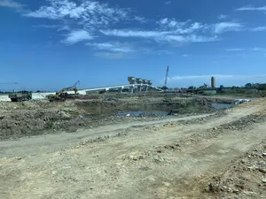 Bối cảnh là một công trường xây dựng quy mô lớn dưới bầu trời xanh và có mây. Ở phía trước là một con đường đất gồ ghề và khu vực đất đã được đào xới, có những vũng nước nhỏ. Nhiều loại máy móc xây dựng đang hoạt động, bao gồm một máy xúc màu vàng, một xe tải ben màu xanh lá cây và một xe tải khác. Ở phía sau, một cây cầu hoặc cầu cạn lớn bằng bê tông đang trong quá trình xây dựng, với các trụ đỡ và các cấu trúc tháp đặc trưng trên mặt cầu. Một cần cẩu cao và một nhà máy xi măng hoặc silo cũng có thể được nhìn thấy ở phía xa. Không có văn bản hoặc biển báo nào có thể đọc được.。（日本語訳）。背景は、青く雲のある空の下の大規模な建設現場です。前景には、でこぼこの未舗装路と掘削された土のエリアがあり、小さな水たまりができています。黄色いショベルカー1台、緑色のダンプトラック1台、そしてもう1台のトラックを含む、多くの種類の建設機械が稼働しています。後方では、橋脚と橋面上の特徴的な塔の構造物を持つ、コンクリート製の大きな橋または高架橋が建設中です。遠くには、高いクレーン1基とセメント工場またはサイロも見えます。読み取れる文字や標識はありません。