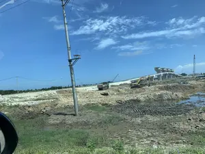 Trên cột điện ở phía trước có một biển báo với biểu tượng tia sét và số "01". Bối cảnh là một công trường xây dựng rộng lớn dưới bầu trời xanh, quang đãng. Một máy xúc màu vàng hiệu CAT và một xe ben màu xanh lá cây đang hoạt động trên bãi đất đá. Phía sau, một cây cầu hoặc cầu cạn lớn bằng bê tông đang được xây dựng, với một cần cẩu tháp ở gần đó. Mặt đất là bùn lầy, đất đã được đào xới và có những vũng nước. Cảnh vật dường như được chụp từ bên trong một chiếc ô tô, vì có thể thấy một phần gương chiếu hậu ở góc dưới bên trái.。（日本語訳）。正面の電柱には、稲妻のシンボルと「01」という数字が書かれた標識があります。背景は、青く澄み渡った空の下に広がる広大な建設現場です。CATブランドの黄色いショベルカーと緑色のダンプトラックが、土砂のある場所で稼働しています。後方では、大きなコンクリート製の橋または高架橋が建設中で、その近くにはタワークレーンがあります。地面はぬかるんでおり、土は掘り起こされ、水たまりができています。