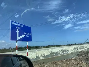 Bức ảnh được chụp từ bên trong một chiếc ô tô, ghi lại cảnh quan ven đường. Đối tượng nổi bật là một biển báo giao thông lớn màu xanh lam. Trên biển báo có ghi chữ trắng chỉ dẫn đường đi: 'UBND XÃ NGA TÂN 4 Km' với mũi tên chỉ thẳng, và 'UBND XÃ ĐA LỘC 3 Km' với mũi tên chỉ rẽ phải. Cảnh vật cho thấy một con đường chạy dọc theo một con kênh hoặc mương nước, có bờ kè được gia cố bằng đá. Bối cảnh xung quanh là vùng nông thôn dưới bầu trời trong xanh, có mây trắng, cột điện và đường dây điện. Ở phía xa có thể nhìn thấy cây cối, một vài ngôi nhà và một chiếc cần cẩu xây dựng. Một phần gương chiếu hậu của ô tô cũng xuất hiện ở góc dưới bên trái.。（日本語訳）。写真は車内から撮影されたもので、道端の風景を記録しています。目立つ対象物は、大きな青い交通標識です。標識には白い文字で道案内が書かれています：まっすぐを指す矢印と共に「NGA TAN村人民委員会 4 Km」、右折を指す矢印と共に「DA LOC村人民委員会 3 Km」。風景は、石で補強された護岸のある運河または水路に沿って走る道を示しています。周囲の背景は、白い雲、電柱、電線のある青空の下の田園地帯です。遠くには木々、いくつかの家、そして建設用のクレーンが見えます。