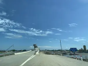 Hình ảnh chụp một công trường xây dựng cầu và cống vào một ngày nắng, trời trong xanh và có mây. Có một biển báo màu xanh lam ghi chữ trắng "CẦU - CỐNG LÈN" và "KM0 + 250". Con đường dẫn lên cầu chưa hoàn thiện, một phần là đường bê tông có vạch kẻ, phần còn lại là đường đất. Cây cầu dẫn đến một công trình cống lớn có tháp điều khiển. Xung quanh là khu vực thi công với đất đá, bãi bùn và các máy móc xây dựng như cần cẩu và trạm trộn bê tông.。（日本語訳）。晴れた日の、青く澄み渡り雲のある空の下で撮影された、橋と水門の建設現場の写真。「CẦU - CỐNG LÈN」および「KM0 + 250」と白字で書かれた青い看板がある。橋へと続く道は未完成で、一部は線が引かれたコンクリートの道路、残りの部分は未舗装の土の道である。その橋は、制御塔のある大きな水門施設へと続いている。周囲は、土や石、ぬかるみ、そしてクレーンやコンクリート混合プラントといった建設機械がある工事エリアである。