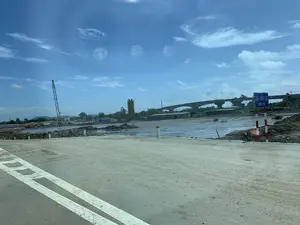 Trên biển báo giao thông màu xanh cắm trên cột sơn sọc đỏ trắng có ghi 'CẦU - CỐNG LÈN' và 'KM0+250'. Bối cảnh là một đại công trường xây dựng cầu vượt sông. Một cây cầu bê tông lớn đang được thi công dở dang ở phía xa. Nhiều máy móc xây dựng có mặt tại hiện trường, bao gồm một cần cẩu tháp lớn ở bên trái và một trạm trộn bê tông. Lòng sông hoặc khu vực xây dựng ngập bùn đất. Phía trước là một con đường đất đang được thi công với vạch kẻ đường màu trắng.。（日本語訳）。赤と白の縞模様に塗られた柱に立てられた青い交通標識には、「CẦU - CỐNG LÈN」と「KM0+250」と書かれている。背景は、川を渡る橋を建設する大規模な工事現場である。遠方では、大きなコンクリート橋が建設途中である。現場には多くの建設機械があり、左側には大きなタワークレーン、そしてコンクリート混合プラントも含まれている。川底または建設エリアは泥でぬかるんでいる。手前には、白い線が引かれた、建設中の未舗装道路がある。