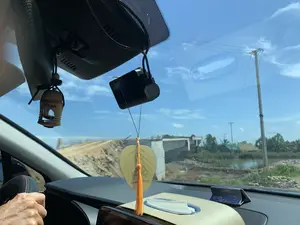 Bức ảnh được chụp từ bên trong một chiếc ô tô, nhìn ra ngoài qua kính chắn gió vào một ngày nắng. Bên trong xe, có một camera hành trình được gắn trên kính. Treo gần gương chiếu hậu là một lọ nước hoa ô tô bằng gỗ có chữ "areon" và một vật trang trí hình chiếc lá bồ đề màu vàng có tua rua màu cam, trên đó có in hình Phật và dòng chữ có thể đọc là "VẠN SỰ AN NHIÊN". Tay của người lái xe đang đặt trên vô lăng. Nhìn ra bên ngoài là một công trường xây dựng, nổi bật là một cây cầu bê tông đang trong quá trình thi công. Xung quanh có các ụ đất, cột điện, cây cối và một vùng nước nhỏ.。（日本語訳）。写真は、ある晴れた日に車内からフロントガラスを通して外を撮影したものです。車内には、ガラスに取り付けられたドライブレコーダーがあります。バックミラーの近くには、「areon」と書かれた木製のカーフレグランスのボトルと、オレンジ色のタッセルが付いた黄色の菩提樹の葉の形をした装飾品が吊るされています。その上には仏陀の絵と「VẠN SỰ AN NHIÊN」と読める文字が印刷されています。運転手の手がハンドルに置かれています。外を見ると建設現場で、目立つのは建設中のコンクリートの橋です。周りには土の山、電柱、木々、そして小さな水域があります。