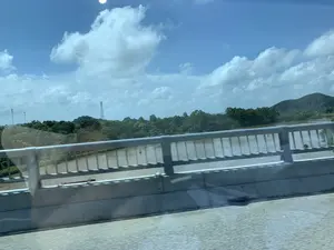 Bức ảnh ghi lại khung cảnh nhìn từ một phương tiện đang di chuyển khi đi qua một cây cầu. Ở tiền cảnh là lan can bê tông và kim loại của cây cầu. Bên dưới là một con sông rộng có dòng nước màu nâu đục. Trên mặt sông có một vài chiếc thuyền nhỏ (ghe) và ít nhất một phao tiêu. Nhìn sang bên kia sông là một vùng cây cối xanh tươi, rậm rạp, phía sau là những ngọn đồi. Có thể thấy một vài cột tháp cao, có thể là cột điện cao thế hoặc cột phát sóng viễn thông. Bầu trời trong xanh, có nhiều mây trắng. Không có văn bản nào có thể đọc được trong ảnh.。（日本語訳）。写真は、橋を渡る際に移動中の乗り物から見た光景を捉えたものです。前景には、橋のコンクリートと金属製の手すりがあります。下には、濁った茶色の水が流れる広い川があります。川面には、数隻の小舟（ゲ）と、少なくとも1つのブイがあります。川の向こう岸には、青々とした木々が密集した地域があり、その後ろには丘があります。いくつかの高い塔が見え、それらは高圧電線の鉄塔か、または電気通信の電波塔である可能性があります。空は青く澄んでおり、白い雲が多く浮かんでいます。写真の中には、読み取れるテキストはありません。