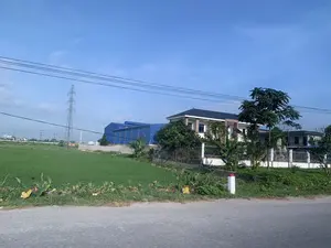 Bức ảnh chụp một khung cảnh ở vùng nông thôn vào một ngày trời quang đãng. Phía trước là một con đường nhựa, bên lề có một cột mốc sơn trắng đỉnh đỏ. Bên trái là một cánh đồng lúa xanh mướt. Bên phải là một ngôi nhà dân kiểu hiện đại, có hàng rào và cây cối xung quanh. Phía sau ngôi nhà là một nhà xưởng lớn lợp tôn màu xanh, trên tường có một logo nhỏ nhưng không thể đọc rõ. Ở phía xa có một cột điện cao thế và các đường dây điện vắt ngang qua bầu trời trong xanh.。（日本語訳）。写真は、晴れた日の田舎の風景を写したものです。手前にはアスファルトの道があり、その脇には上部が赤く塗られた白い標柱があります。左側には青々とした水田が広がっています。右側には現代的なスタイルの民家があり、その周りには柵や木々があります。遠くには高圧電線の鉄塔があり、電線が青空を横切っています。