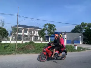 Trên biển hiệu của tòa nhà có ghi 'CÔNG TY TNHH MINH THUYẾT TH' và 'TỔNG KHO LẠNH'. Trên cánh cổng màu xanh có chữ viết 'KÉO RA'. Một cột mốc bên đường ghi 'H8' và '68'. Bối cảnh là một con đường nhựa ở khu vực ngoại ô hoặc nông thôn vào một ngày nắng đẹp, trời trong xanh. Hai người, một nam và một nữ, đang đi trên một chiếc xe máy tay ga màu đỏ. Người đàn ông lái xe mặc đồ đen, đội mũ bảo hiểm màu cam. Người phụ nữ ngồi sau mặc áo màu hồng, đội mũ bảo hiểm màu vàng. Ở hậu cảnh, có một ngôi nhà hai tầng màu trắng, mái đen, được bao quanh bởi hàng rào. Bên cạnh là tòa nhà của một công ty với cổng màu xanh, được xác định là một tổng kho lạnh. Dọc bên đường có cây cối, cỏ và cột điện.。（日本語訳）。建物の看板には「MINH THUYET TH有限会社」と「総合冷蔵倉庫」と書かれている。青い門扉には「引く」という文字が書かれている。道端のキロポストには「H8」と「68」と記されている。背景は、晴れた美しい日の郊外または田舎の地域にあるアスファルトの道で、空は青く澄んでいる。男性一人と女性一人の二人が、赤いスクーターに乗っている。運転している男性は黒い服を着て、オレンジ色のヘルメットをかぶっている。後ろに乗っている女性はピンク色の服を着て、黄色いヘルメットをかぶっている。背景には、黒い屋根の白い二階建ての家があり、柵で囲まれている。その隣には青い門のある会社の建物があり、総合冷蔵倉庫だとわかる。道沿いには木々、草、電柱がある。