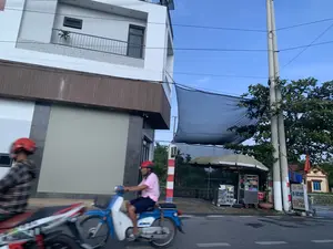 Trên một con đường nhựa, có hai người đang đi xe máy. Người đi trước bị mờ do chuyển động, người đi sau là một người đàn ông đội mũ bảo hiểm màu đỏ, mặc áo phông hồng, đang lái một chiếc xe máy màu xanh. Bên trái là một ngôi nhà phố nhiều tầng, hiện đại, màu trắng, có ban công và cửa cuốn ở tầng trệt đang đóng kín. Bên phải đường là một quán nước vỉa hè, có một xe đẩy bán hàng, vài chiếc ô lớn và một tấm lưới che nắng màu đen được giăng lên. Một tấm biển quảng cáo dựng gần đó có ghi chữ: 'GIẢI KHÁT', 'BIA - NƯỚC NGỌT', 'NƯỚC MÍA'. Phía sau quán nước là cây cối, cột điện và một phần của một ngôi nhà khác.。（日本語訳）。アスファルトの道路の上を、二人がバイクで走っています。前の人は動きでぼやけており、後ろの人は赤いヘルメットをかぶり、ピンクのTシャツを着た男性で、青いバイクを運転しています。左側には、多層階でモダンな白いタウンハウスがあり、バルコニーと、1階には固く閉ざされたシャッターがあります。道路の右側には歩道のドリンクスタンドがあり、屋台、いくつかの大きな傘、そして張られた黒い日よけネットがあります。近くに立てられた看板には、「GIẢI KHÁT」、「BIA - NƯỚC NGỌT」、「NƯỚC MÍA」という文字が書かれています。ドリンクスタンドの後ろには、木々、電柱、そして別の家の一部があります。