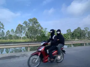 Bức ảnh chụp cảnh hai người đang đi trên một chiếc xe máy màu đỏ trên một con đường nhựa. Cả hai người đều mặc đồ tối màu, áo khoác có mũ và đeo khẩu trang che kín mặt. Họ đang di chuyển trên một con đường có thể là cầu hoặc đường ven kênh, vì có lan can và một con kênh hoặc sông ở bên cạnh. Phía bên kia bờ kênh là hàng cây xanh dưới bầu trời xanh nhạt có mây. Hình ảnh bị mờ do chuyển động, cho thấy chiếc xe máy đang chạy.。（日本語訳）。赤いバイクに乗ってアスファルトの道路を走っている2人の光景を撮った写真です。2人とも暗い色の服、フード付きのジャケットを着ており、顔を覆うマスクをしています。彼らは、橋または運河沿いの道路と思われる道を進んでいます。なぜなら、欄干があり、その隣には運河か川があるからです。運河の対岸には、雲のある薄青い空の下に緑の木々が並んでいます。画像は動きによってぼやけており、バイクが走行中であることを示しています。