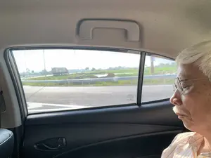 Bức ảnh được chụp từ ghế sau của một chiếc ô tô đang di chuyển. Trong ảnh, một người đàn ông lớn tuổi có mái tóc bạc, đeo kính, đang nhắm mắt nghỉ ngơi. Nhìn qua cửa sổ xe, có thể thấy khung cảnh vùng nông thôn với những cánh đồng và ao hồ. Chiếc xe đang chạy trên một con đường có lan can bảo vệ. Ở phía xa có một chiếc xe tải. Trên cửa kính xe có một số dòng chữ thông số kỹ thuật, trong đó có chữ 'TOYOTA' bị ngược.。（日本語訳）。写真は、移動中の車の後部座席から撮影されたものです。写真には、白髪で眼鏡をかけた年配の男性が、目を閉じて休んでいます。車の窓からは、田んぼや池のある田園風景が見えます。車はガードレールのある道路を走っています。遠くにはトラックが1台います。車の窓ガラスにはいくつかの仕様に関する文字があり、その中には逆さまになった「TOYOTA」の文字があります。