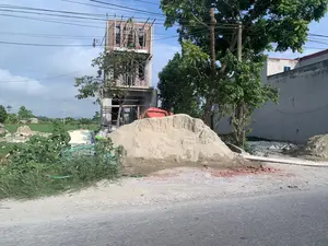 背景は、ある農村地域の道路沿いにある建設現場です。赤レンガの壁とコンクリートの骨組みを持つ、多層階建ての家が建設中です。最上階は竹製の足場で囲まれ、作業員が働いています。手前の道端には、大きな砂の山、赤いコンクリートミキサー、そして割れたレンガの破片があります。周囲には木々や、多くの電線が張られた電柱があります。遠くには緑の畑と、他の家が数軒あります