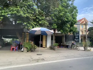 Trên một tấm biển hiệu có ghi: 'L/H: 0987 438 159', 'NAIL MI SPA', 'Phạm Thùy Linh'. Đây là khung cảnh ven đường với nhiều cây xanh. Bên lề đường có các cửa hàng, trong đó có một tiệm làm đẹp. Một người đội nón lá đang đứng cạnh một chiếc xe đạp. Gần đó có một chiếc ô lớn màu xanh trắng, một xe đẩy chở đầy trái cây trông giống như dừa, và một vài bộ bàn ghế nhựa. Phía sau là các ngôi nhà, bao gồm một ngôi nhà cao tầng có mái ngói đỏ.。（日本語訳）。ある看板に「L/H: 0987 438 159」「NAIL MI SPA」「Phạm Thùy Linh」と書かれています。これはたくさんの緑の木々がある道端の風景です。道端には店があり、その中には美容サロンが1軒あります。ノンラーをかぶった人が自転車の隣に立っています。後ろには家々があり、その中には赤い瓦屋根の多層階の家も含まれています。