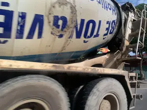 Hình ảnh chụp cận cảnh một chiếc xe bồn trộn bê tông đang di chuyển trên đường. Do ảnh được chụp khi đang chuyển động nên có phần bị mờ. Thân xe và các bánh xe lớn dính đầy bùn đất. Trên bồn trộn màu trắng có dòng chữ lớn màu xanh lam, một phần có thể đọc được là 'BÊ TÔNG VIỆT' và 'HOTLINE' cùng với số '38'. Chữ 'BÊ TÔNG' được viết ngược, đây là một đặc điểm thường thấy trên các loại xe này. Phía sau xe là cây cối và một phần mái nhà ven đường.。（日本語訳）。道路を走行中のコンクリートミキサー車を接写した画像です。動いているときに撮影されたため、写真の一部はぼやけています。車体と大きな車輪は泥で汚れています。白いミキサータンクには大きな青い文字があり、その一部は「BÊ TÔNG VIỆT」と「HOTLINE」、そして数字の「38」と読み取れます。「BÊ TÔNG」の文字は逆さに書かれており、これはこの種の車両によく見られる特徴です。車両の後ろには木々と、道端の家屋の屋根の一部が見えます。