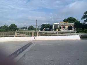 Bức ảnh chụp khung cảnh ven đường từ trên một cây cầu. Ở phía bên kia đường là một tòa nhà kinh doanh dịch vụ ăn uống. Trên mái nhà có biển hiệu lớn màu vàng ghi "Ẩm Thực Cây Đa", bên dưới là dòng chữ "Lẩu - Nướng - Cơm Quê". Một biển hiệu khác phía dưới quảng cáo "BÚN BÒ HUẾ - LẨU ĐỒNG QUÊ - ĐUÔI BÒ - BIA HƠI". Bên cạnh đó là quán "Cafe Cây Đa" bán "TRÀ CHANH - TRÀ SỮA - KEM - CHÈ - MỲ CAY". Ngoài ra còn có một biển hiệu nhỏ màu đỏ ghi "BÚN BÒ HUẾ". Khung cảnh cho thấy một cây cầu có mặt đường nhựa, lan can kim loại và dải phân cách bê tông. Xung quanh là cây cối, một vài ngôi nhà khác và đường dây điện trên bầu trời nhiều mây. Bên dưới cầu có thể là một con sông hoặc kênh nhỏ.。（日本語訳）。橋の上から道端の風景を撮った写真です。道の向こう側には、飲食サービスを営む建物があります。屋根の上には「Ẩm Thực Cây Đa」と書かれた黄色い大きな看板があり、その下には「Lẩu - Nướng - Cơm Quê」という文字があります。その下にある別の看板には「BÚN BÒ HUẾ - LẨU ĐỒNG QUÊ - ĐUÔI BÒ - BIA HƠI」と広告されています。その隣には「TRÀ CHANH - TRÀ SỮA - KEM - CHÈ - MỲ CAY」を販売する「Cafe Cây Đa」があります。さらに、「BÚN BÒ HUẾ」と書かれた小さな赤い看板もあります。周りには木々、他のいくつかの家、そして曇り空に電線が見えます。橋の下は川か小さな水路かもしれません。