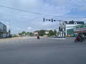 Bản ghi các thông tin văn bản trên các biển hiệu:。- Biển hiệu lớn màu xanh lá cây của cửa hàng kính thuốc: "KÍNH THUỐC SÀI GÒN", "Y BÁC SĨ CHUYÊN KHOA KHÚC XẠ", "CẬN - VIỄN - LOẠN - HAI TRÒNG - ĐA TRÒNG", và địa chỉ "KHU 4 - NGÃ TƯ THỊ TRẤN QUẢN LÀO - YÊN ĐỊNH - THANH HÓA". Một biển hiệu khác ghi: "CHUYÊN NGÀNH KÍNH ĐỘ", "CẬN, VIỄN, LOẠN, ĐA TRÒNG, 2 TRÒNG, ÁP TRÒNG", "ĐO MẮT, CẮT KÍNH & TƯ VẤN THỊ LỰC CHUYÊN NGHIỆP".。- Băng rôn đỏ: "CHÀO MỪNG ĐẠI LỄ 30/4 - 1/5".。- Biển hiệu quán cà phê: "HÙNG PHAT COFFEE & TEA".。- Biển quảng cáo: "LG OLED".。- Biển hiệu màu hồng trên tòa nhà trắng: "CHO THUÊ CĂN HỘ" và số điện thoại liên hệ không đọc rõ.。- Biển hiệu nhỏ màu xanh lá: "CẬN VIỄN LOẠN".。Mô tả khung cảnh và hoạt động:。Bức ảnh chụp một ngã tư đường phố tại khu vực thị trấn Quản Lào, Yên Định, Thanh Hóa. Bầu trời có mây. Nhiều người đang điều khiển xe máy tham gia giao thông tại ngã tư. Bên phải là một tòa nhà lớn màu trắng theo kiến trúc hiện đại và cửa hàng "Kính thuốc Sài Gòn". Bên trái là một khu đất trống đang được thi công, có một chiếc máy xúc màu vàng và một biển quảng cáo lớn. Phía xa hơn là một quán cà phê. Hệ thống đèn tín hiệu giao thông và dây điện được giăng mắc phía trên đường. Có các rào chắn tạm thời màu xanh-trắng được đặt ở giữa đường để phân luồng giao thông.。（日本語訳）。看板のテキスト情報の記録：。- 眼鏡店の大きな緑色の看板：「サイゴン眼鏡」、「屈折専門医」、「近視 - 遠視 - 乱視 - 二重焦点 - 多焦点」、および住所「第4区 - クアンラオ町交差点 - イエンディン - タインホア」。別の看板には、「度付き眼鏡専門」、「近視、遠視、乱視、多焦点、二重焦点、コンタクトレンズ」、「専門的な視力測定、眼鏡作成、視力相談」と書かれている。- 赤い横断幕：「4月30日～5月1日の祝日を歓迎します」。- カフェの看板：「フンファット・コーヒー＆ティー」。- 広告看板：「LG OLED」。- 白い建物にあるピンク色の看板：「アパート賃貸」、および連絡先の電話番号は読み取れず。- 小さな緑色の看板：「近視 遠視 乱視」。風景と活動の説明：。写真はタインホア省イエンディン県クアンラオ町の交差点を撮影したものです。空は曇っています。多くの人々が交差点でバイクを運転して交通に参加しています。右側には現代的な建築様式の大きな白い建物と「サイゴン眼鏡」店があります。左側には工事中の空き地があり、黄色いショベルカーと大きな広告看板があります。その先にはカフェがあります。道路の上には信号機システムと電線が張り巡らされています。交通を整理するために、青と白の仮設の柵が道路の中央に設置されています。